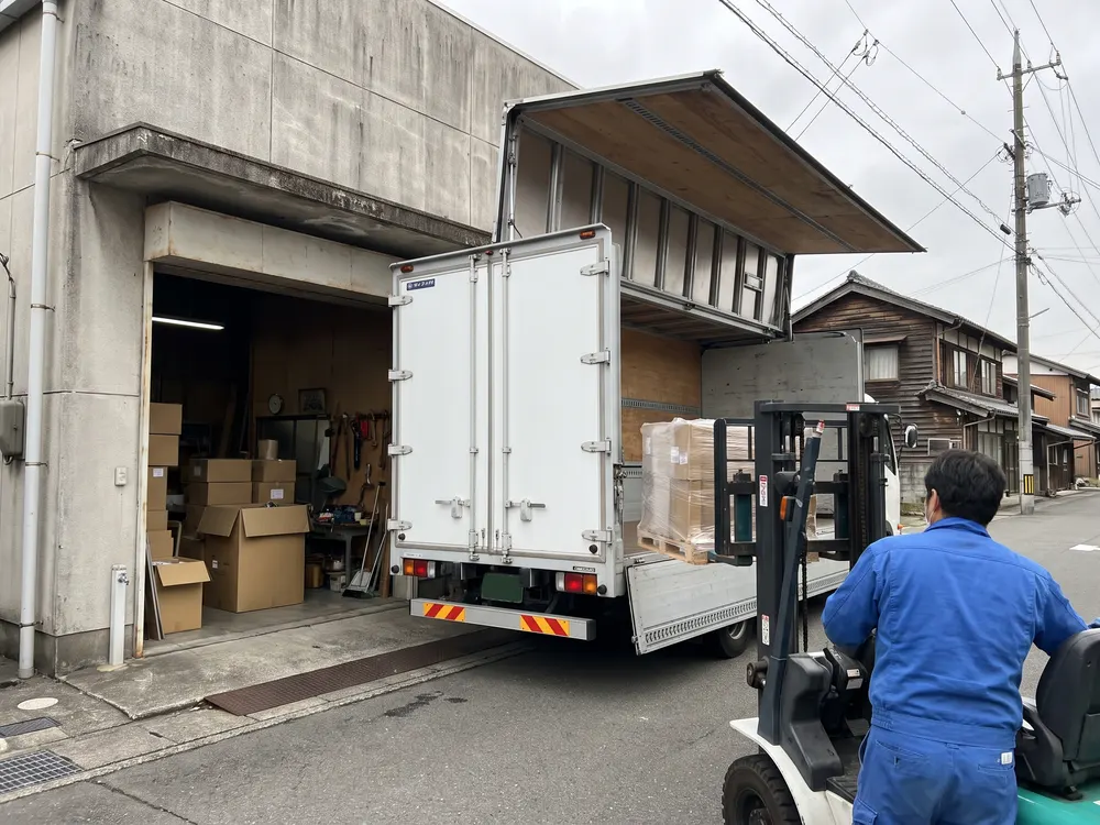 横の扉を開けたトラックにパレットを積むフォークリフト
